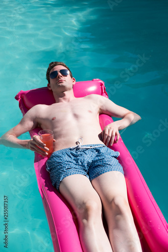 Fit Caucasian man in sunglasses floating on a pink raft in a swimming pool holding a drink. Man on vacation, enjoying the sun in an outdoor swimming pool, nice blue water, thin, good looking young man