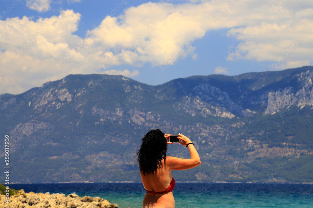 Beautiful brunette girl with long hair and red swimsuit makes selfie on the beach against the backdrop of the picturesque mountains