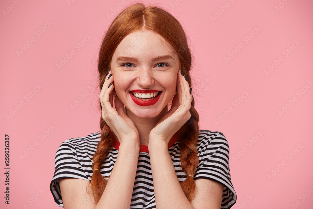 Close up of gorgeous pretty red-haired girl with two braids keeps hands near her face and smiling lively with red lips, showing white healthy teeth, isolated on pink background Close up of gorgeous pretty red-haired girl with two braids keeps hands near her face and smiling lively with red lips, showing white healthy teeth, isolated on pink background