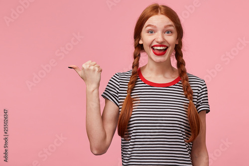 Overflowing with positive emotions red-haired girl with two braids pointing with thumb to the left side on blank copy space, openes mouth widely in excitement, with red lips, isolated on pink wall photo