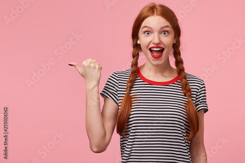 Excited red-haired girl with two braids pointing with thumb to the left side on blank copy space, openes mouth widely in surprise, draws your attention, with red lips, isolated on pink wall photo