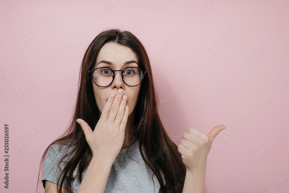 Attractive young female in eyeglasses covers mouth with palm, attracts your attention at copy space aside, dressed in gray t-shirt, stands against pink wall. People, reaction and emotions concept Attractive young female in eyeglasses covers mouth with palm, attracts your attention at copy space aside, dressed in gray t-shirt, stands against pink wall. People, reaction and emotions concept
