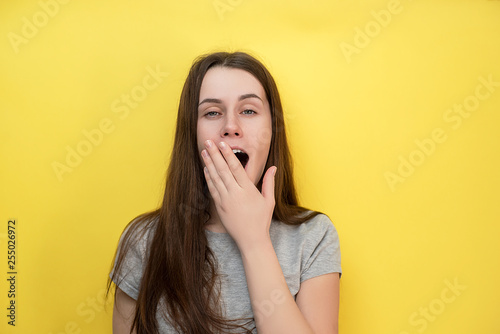 Shot of sleepy attractive caucasian young woman , dressed in gray T-shirt, feeling tired after night without sleep, yawning, covering opened mouth with palm, models against yellow background