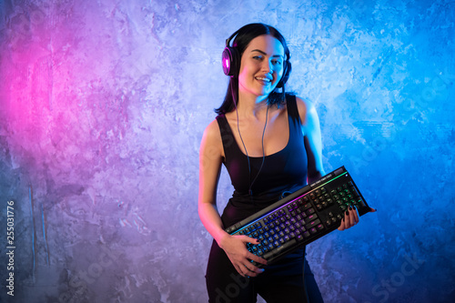 Portrait of the Beautiful Young Pro Gamer Girl standing with a gaming keyboard and headset and Looks into Camera. Attractive Geek Girl Player Wearing Glasses in the Room Lit by Neon Lights.