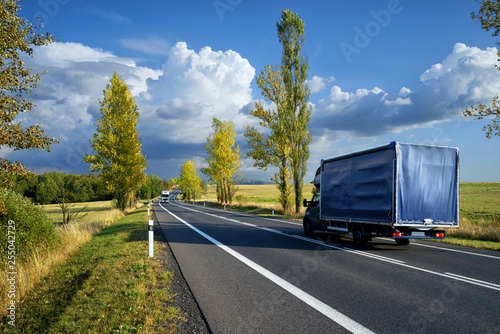 Obraz na płótnie Blue delivery van and white truck driving on the asphalt road between line of trees in autumn landscape w salonie