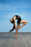 A young girl in a black gymnastic swimsuit gymnast makes an exercise standing backward and bent knee on a blue backgroun
