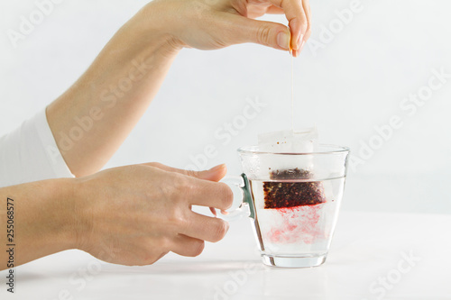 Close-up of a woman hand dipping a sachet of Hibiscus Tea in a glass cup full of water with beautiful red-colored effects in the transparency of water