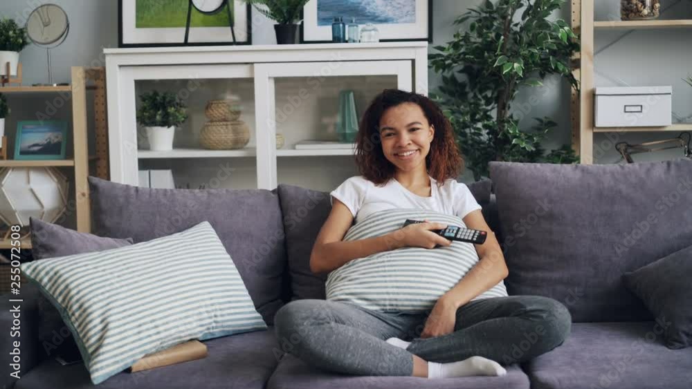 Zoom-in of pretty African American student watching funny comedy on TV and laughing sitting on sofa and holding remote control. Television and emotions concept. Zoom-in of pretty African American student watching funny comedy on TV and laughing sitting on sofa and holding remote control. Television and emotions concept.