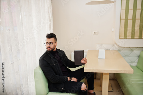 Arab man wear on black jeans jacket and eyeglasses sitting in cafe, read book. Stylish and fashionable arabian model guy.