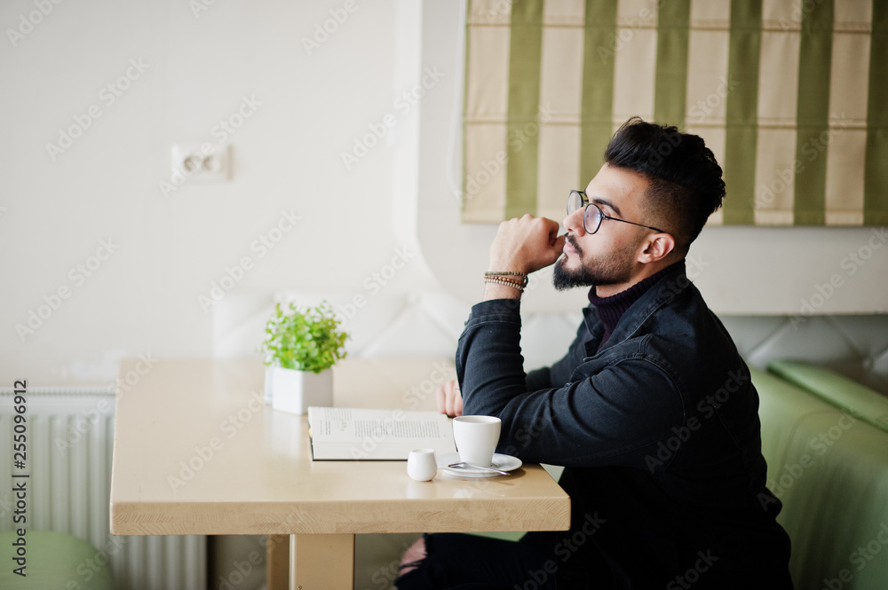 Arab man wear on black jeans jacket and eyeglasses sitting in cafe, read book and drink coffee. Stylish and fashionable arabian model guy. Arab man wear on black jeans jacket and eyeglasses sitting in cafe, read book and drink coffee. Stylish and fashionable arabian model guy.