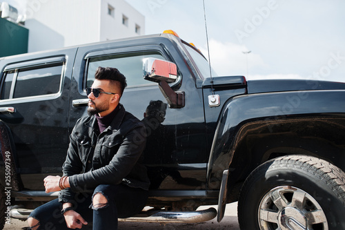 Fashion rich beard Arab man wear on black jeans jacket and sunglasses posed against big black suv car. Stylish, succesful and fashionable arabian model guy. © AS Photo Family