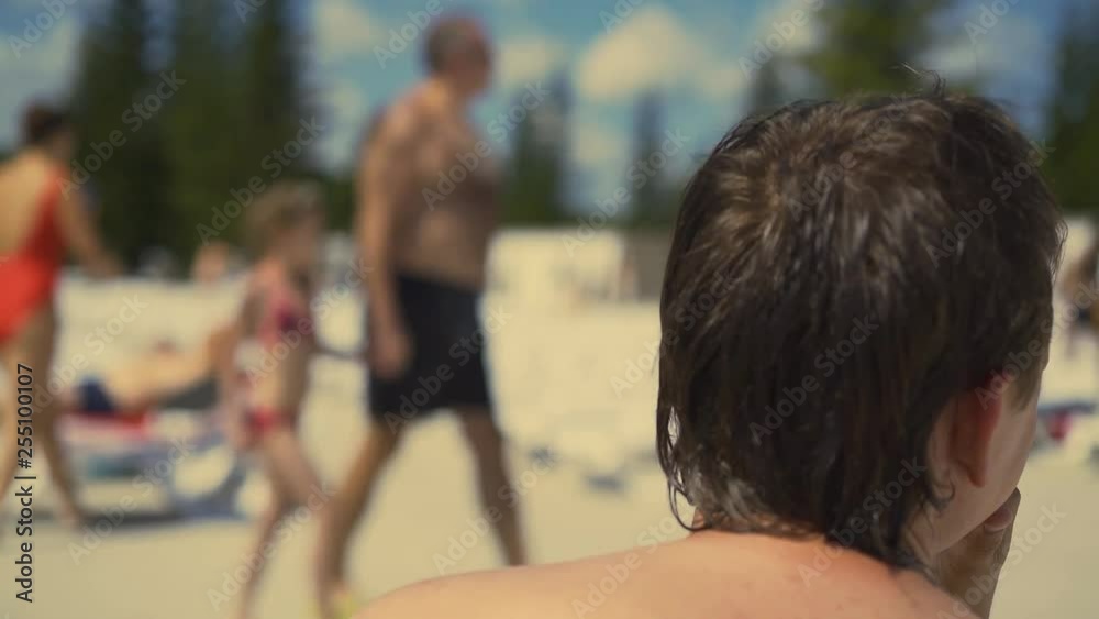 Back view, Close-up: Young man is sitting on the beach on sun lounger. Looking for somebody with his eyes, then, makes sun visor with his hand, finds and runs to the side. Back view, Close-up: Young man is sitting on the beach on sun lounger. Looking for somebody with his eyes, then, makes sun visor with his hand, finds and runs to the side.