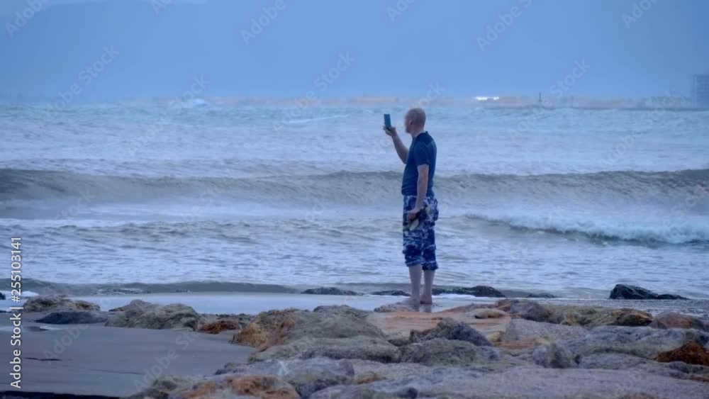 Resting tourist stands on the shore of a stormy sea with big waves and shoots the ocean on a smartphone Resting tourist stands on the shore of a stormy sea with big waves and shoots the ocean on a smartphone