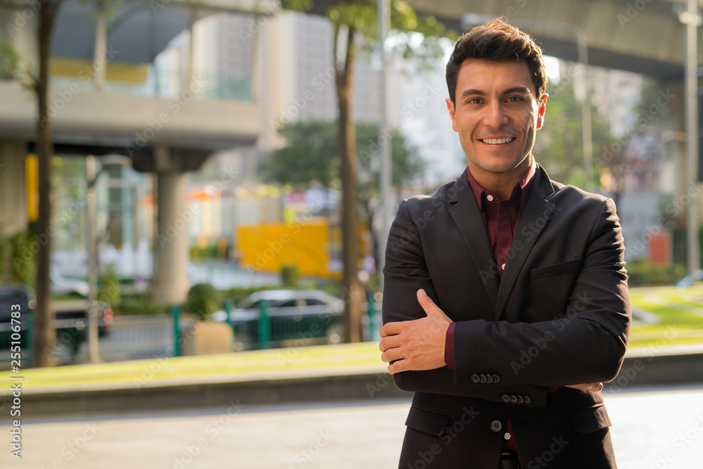 Fototapeta premium Young happy Hispanic businessman smiling with arms crossed outdoors