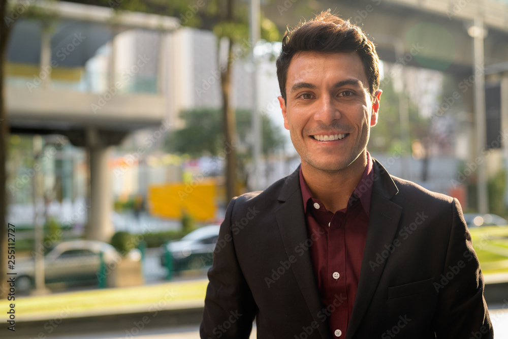 Face of happy young handsome Hispanic businessman smiling in the city - obrazy, fototapety, plakaty 