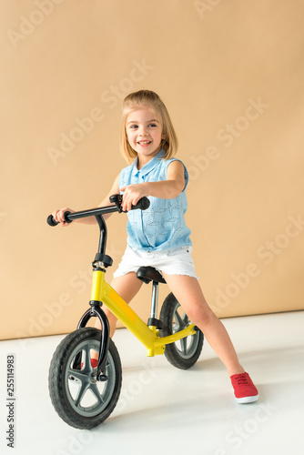 smiling kid in shirt and shorts riding bicycle on beige background