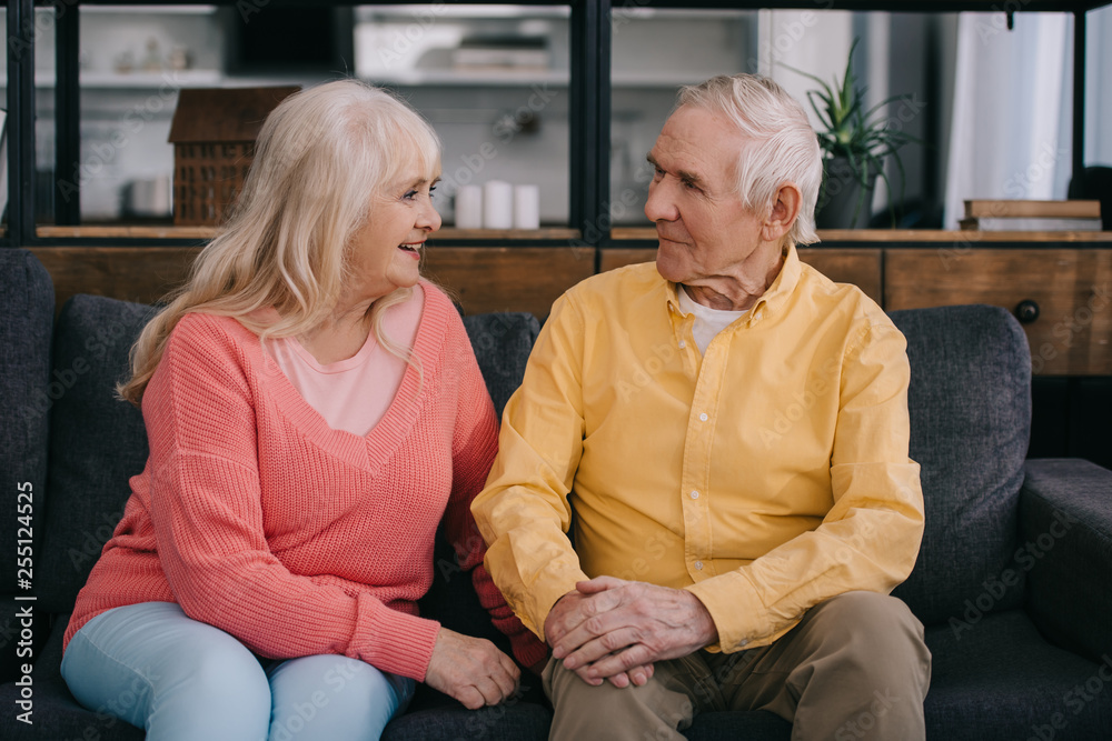 senior couple in casual clothes sitting on couch and looking at each other in living room