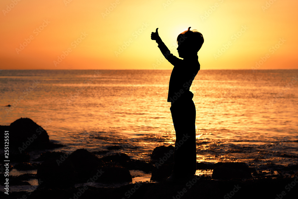contour of a boy with thumbs raised at sunrise sunset on the seashore of the ocean orange sky orange sea ocean
