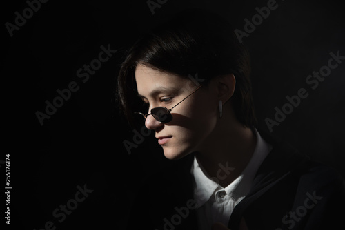 Serious, beautiful girl with short hair sitting in sunglasses under bright light on dark background