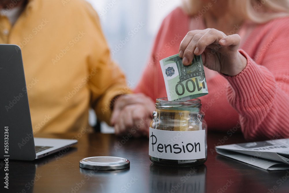Fototapeta premium cropped view of senior couple putting money in glass jar with 'pension' word