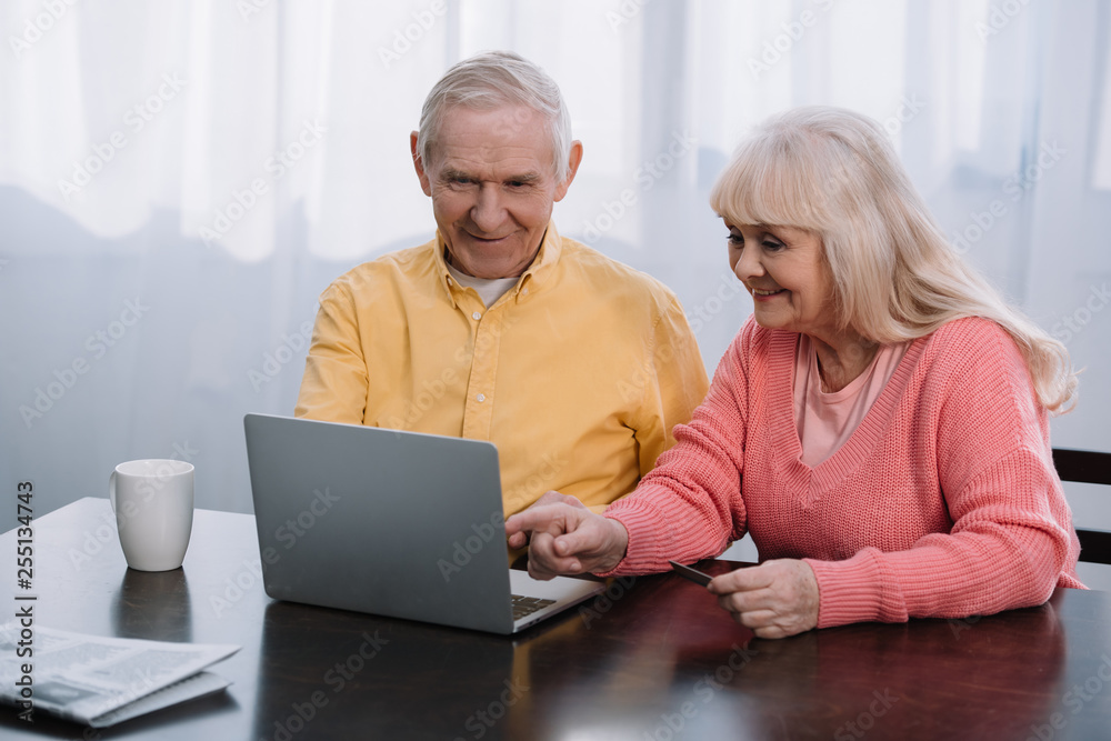 smiling senior couple in casual clothes using laptop while sitting at table at home