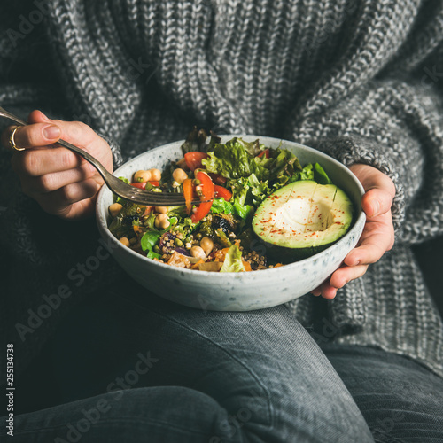 Healthy vegetarian dinner. Woman in grey jeans and sweater eating fresh salad, avocado, grains, beans, roasted vegetables from Buddha bowl, square crop. Superfood, clean eating, dieting food concept photo