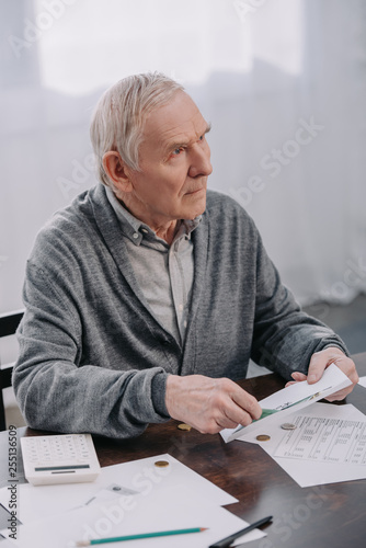 senior man sitting at table with paperwork and holding envelope while counting money
