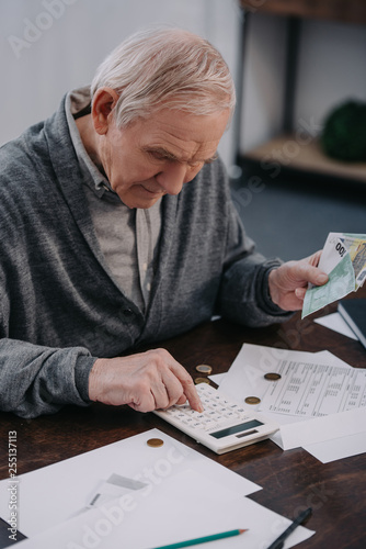 senior man sitting at table with paperwork and using calculator while counting money at home