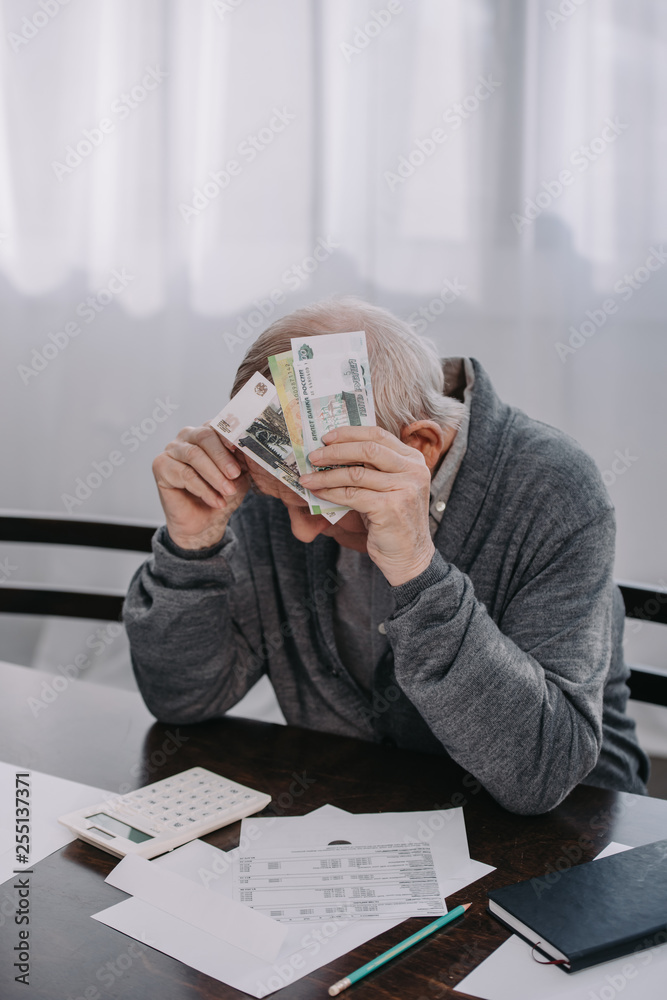 stressed senior man sitting at table with paperwork and holding money at home stressed senior man sitting at table with paperwork and holding money at home