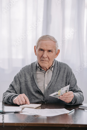 senior man sitting at table with paperwork, looking at camera and using calculator while counting money