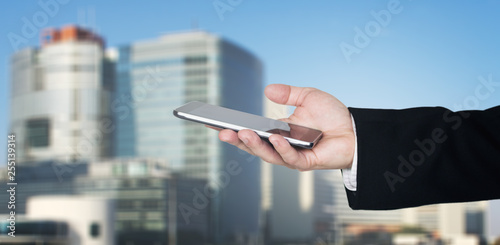 Businessmans Hand Holding Smartphone With Business City and Corporate Buildings In Background