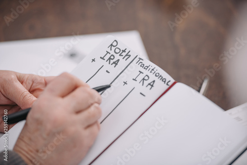 cropped view of senior woman writing in notebook with roth ira and traditional ira words photo