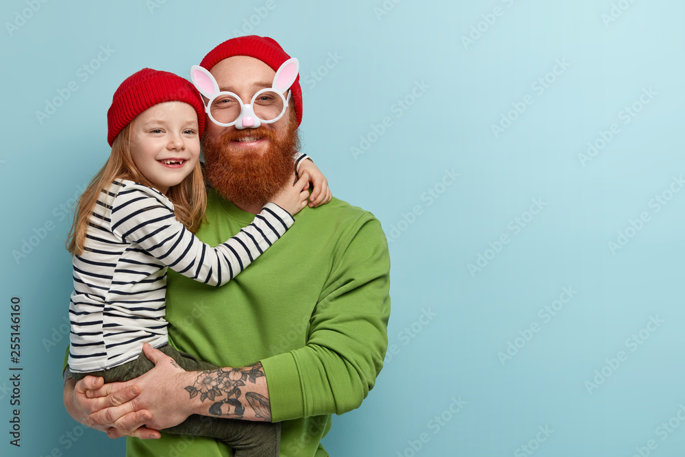 Indoor shot of funny bearded dad wears comic bunny spectacles, plays with little daughter, prepare for Easter party. Small girl in red hat, striped sweater glad to be on fathers hands, embraces him Indoor shot of funny bearded dad wears comic bunny spectacles, plays with little daughter, prepare for Easter party. Small girl in red hat, striped sweater glad to be on fathers hands, embraces him