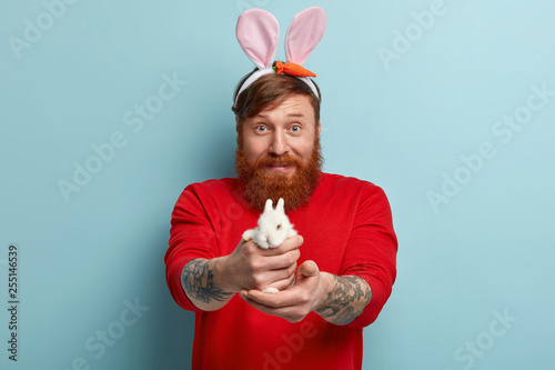 Funny red haired man in casual clothes, suggests to buy small decorative white bunny, wears long rabbits ears, defends rights of animals, has tattoos on arms, ready to celebrate Easter holiday photo