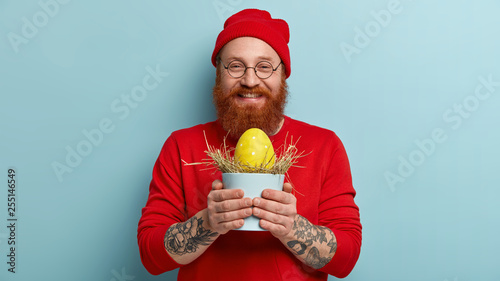 Look at my first decorated Easter egg. Smiling redhead satisfied man carries pot with hay and yellow egg, wears red clothes, ready to hide in garden for children, isolated over blue background photo