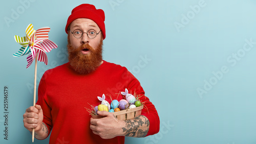 Easter celebration concept. Stupefied man with foxy stubble, carries basket with dyed eggs on hay, holds paper windmill, shocked to find out religious rules during fixed feast, models indoor