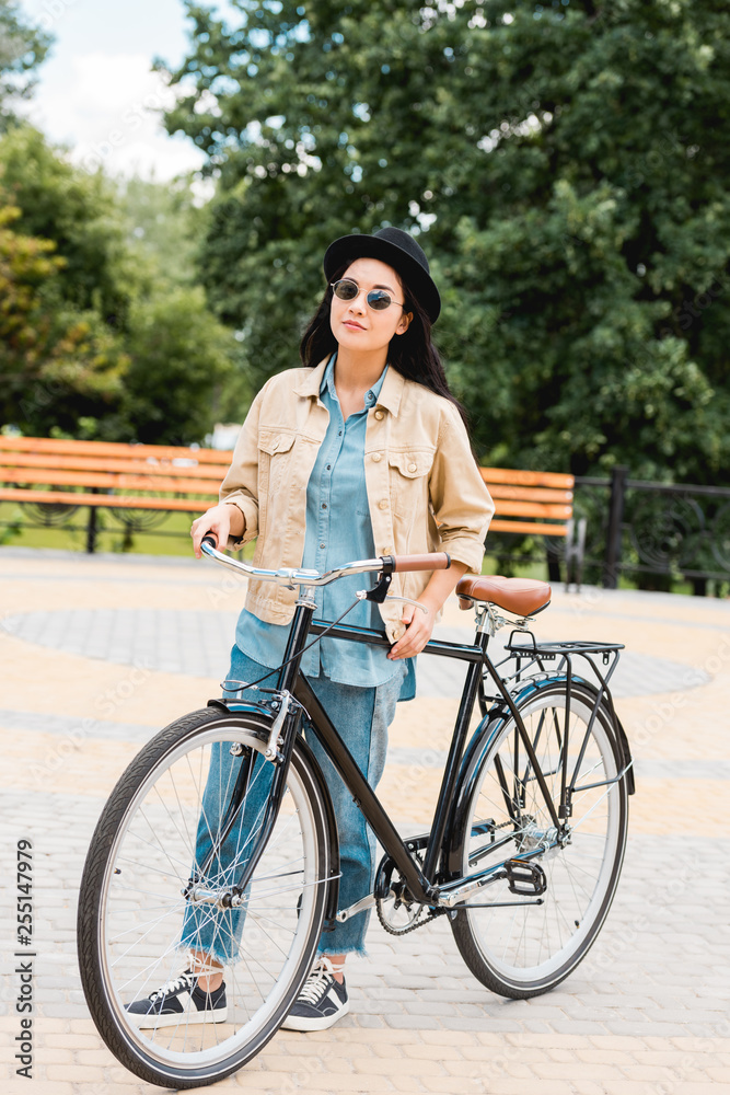 cheerful girl in sunglasses and hat standing with bicycle in park