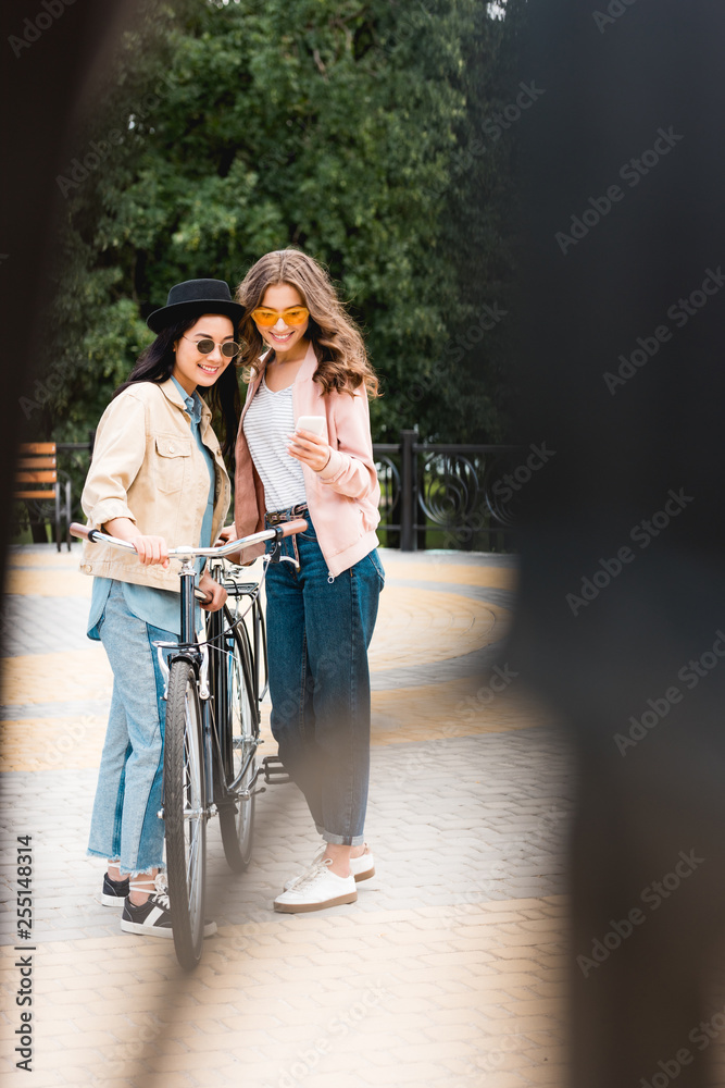 selective focus of cheerful girls in sunglasses smiling while looking at smartphone and standing near bike in park