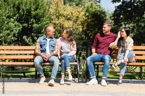 Canvas Print cheerful multicultural friends sitting on benches in park