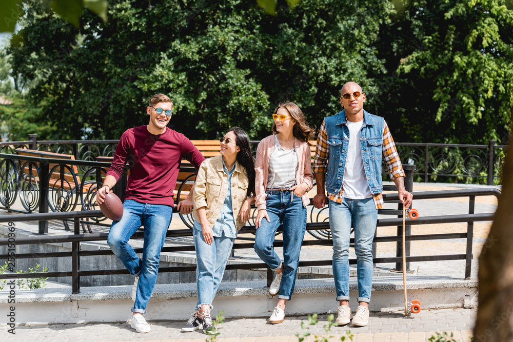 Fototapeta premium Cheerful multicultural young men standing with longboard and American football near girls