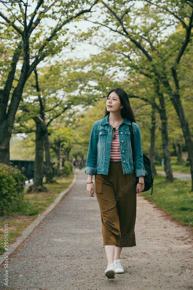 Full length chinese woman backpacker walking in spring park. elegant young college girl student in outdoor in scenic park under big trees on trail on sunny day afternoon going home after course. Full length chinese woman backpacker walking in spring park. elegant young college girl student in outdoor in scenic park under big trees on trail on sunny day afternoon going home after course.