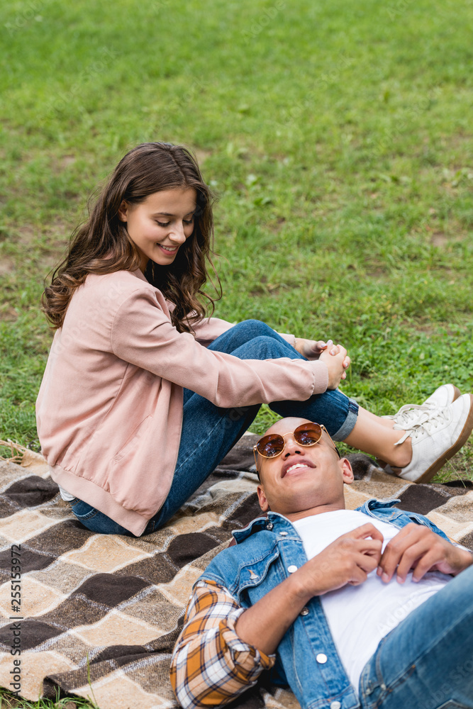 attractive girl looking at african american boyfriend lying on blanket in park attractive girl looking at african american boyfriend lying on blanket in park
