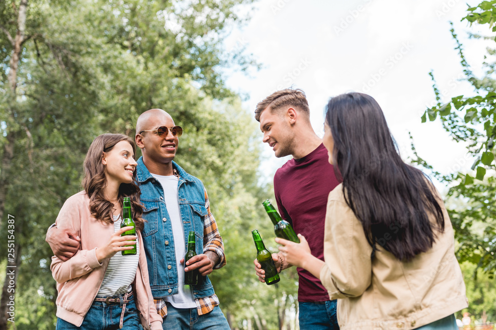 cheerful multicultural friends holding bottles with beer and talking in park