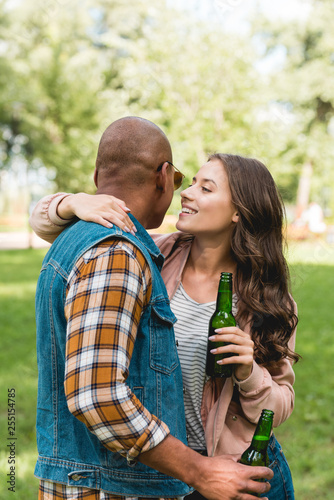 cheerful girl whispering in ear of african american boyfriend while holding bottle