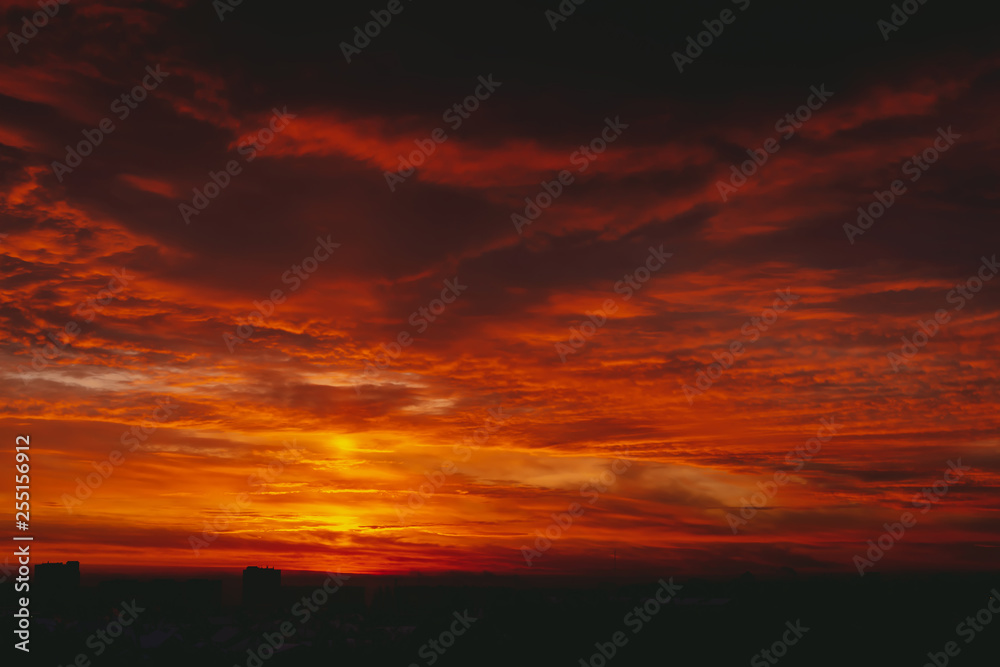 Cityscape with fiery blood vampire dawn. Amazing warm dramatic cloudy sky above dark silhouettes of city buildings. Orange sunlight. Atmospheric background of sunrise in overcast weather. Copy space.