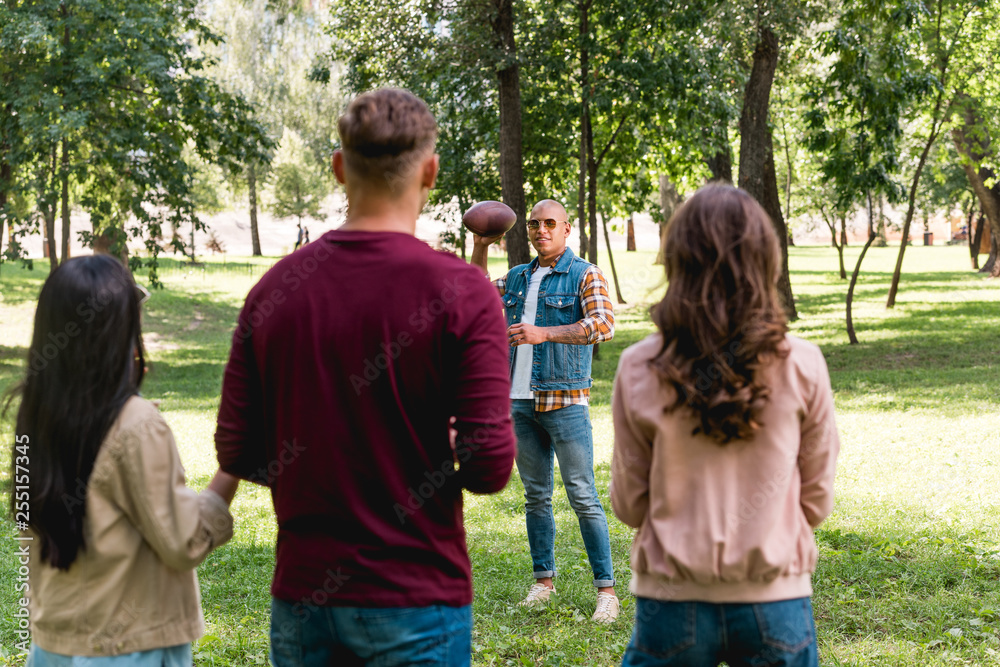 Fototapeta premium back view of friends looking at african american man holding american football in park