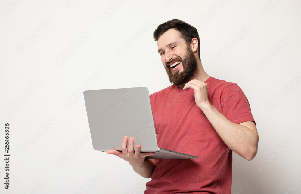 Portrait of a joyful young man in red t-shirt looking at laptop computer and celebrating isolated over grey background 