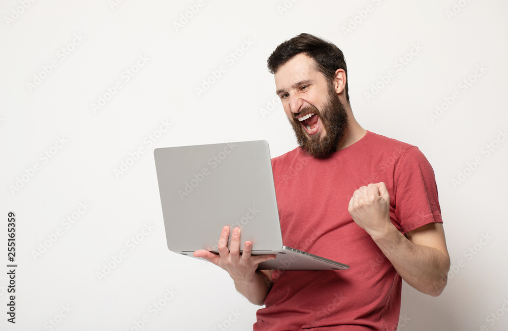 Portrait of a joyful young man in red t-shirt looking at laptop computer and celebrating isolated over grey background Portrait of a joyful young man in red t-shirt looking at laptop computer and celebrating isolated over grey background