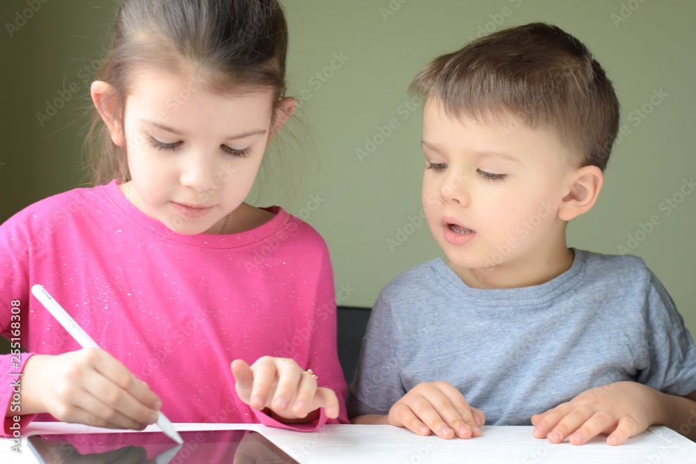 White caucasian toddler boy and beautiful girl drawing together on tablet screen. Portrait of a happiness little brother and sister. Children play a computer game on the tablet White caucasian toddler boy and beautiful girl drawing together on tablet screen. Portrait of a happiness little brother and sister. Children play a computer game on the tablet