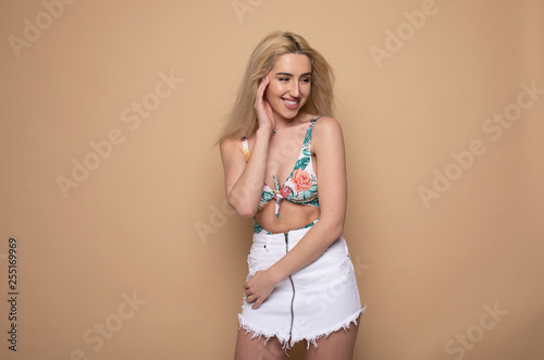 Positive attractive blonde young model wears swimsuit and white skirt being happy to receive good news. Joyful woman rejoices weekends, relaxing indoors, isolated against beige background.